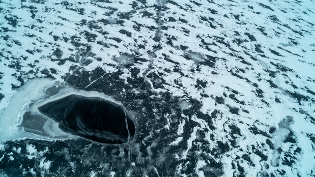 A striking top-down drone photograph looking straight down at a partially frozen winter lake, featuring a dramatic deep black open ice hole surrounded by complex white snow and slush patterns.