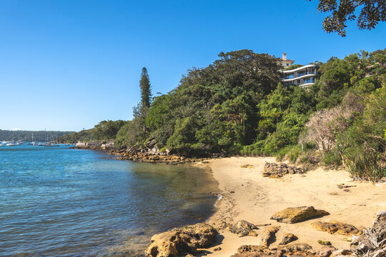 Collins Beach, Sydney&rsquo;s 80km Bondi to Manly coastal walk. This eye-level perspective captures a secluded sandy beach scattered with large, smooth rocks along the water's edge. A modern, multi-story lu