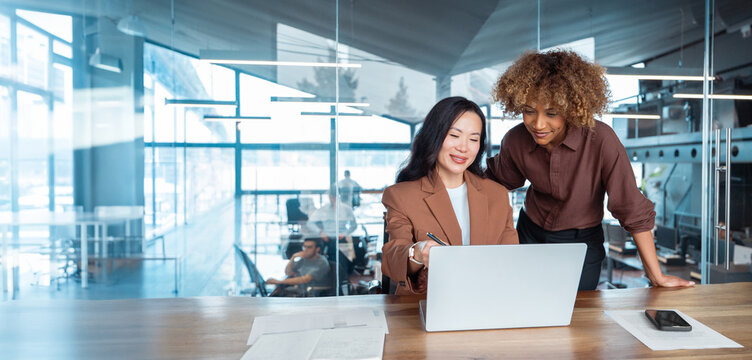 Two businesswomen collaborate at laptop in office, one standing and mentoring at desk. Professional teamwork and business training. Multicultural team working together on project. Banner, copy space