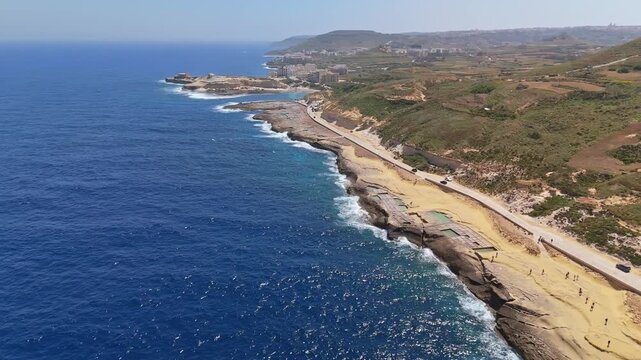 Panoramic drone view of Xwejni salt pans in Żebbuġ, Gozo, where ancient geometric basins edge a rugged Mediterranean shore beneath clear skies and shimmering blue sea.