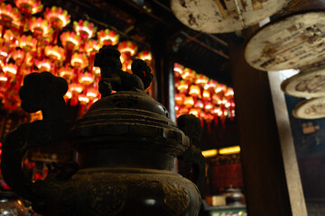 An ornate bronze censer in the foreground of the Tin Hau Temple in Causeway Bay, Hong Kong, with a blurred ceiling of glowing lotus lanterns and an ash-covered incense coil catcher to the right © Vidu Gunaratna