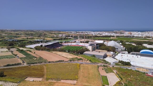 In Ta' Qali, Malta, the national football stadium sits within a suburban landscape of parkland, fields, and surrounding infrastructure seen in a clear-sky bird's-eye view.