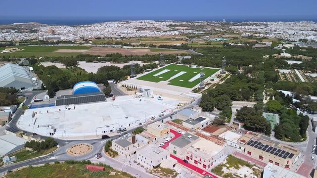 At Ta' Qali National Park in Malta, an aerial view captures the urban park's lawns, trees, walking paths, and surrounding event and craft venues within the Mediterranean landscape.