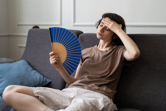 Woman at home suffering from migraine and overheating on sofa in living room waving hand paper fan and touching head. Young female on couch with sick expression, overwhelmed by summer heat or fever.
