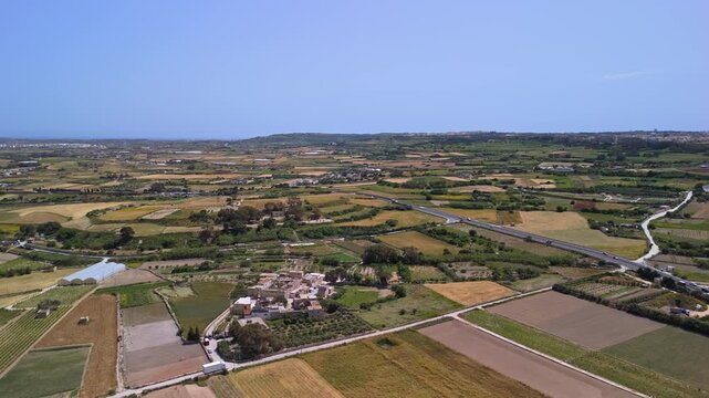 In Ta' Qali, Malta, a drone captures agricultural fields and scattered farmhouses surrounding the rural settlement, set against Mediterranean greenery under a clear daytime sky.