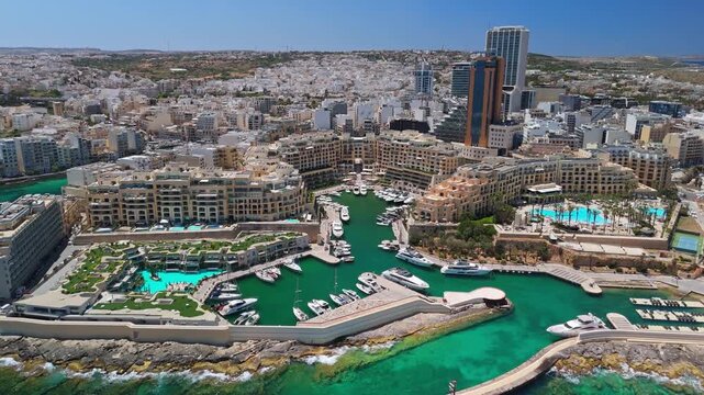In St Julian's, Malta, the cityscape featuring Portomaso Tower, Mercury Tower, and the marina with yachts lines the Mediterranean coastline under clear conditions.