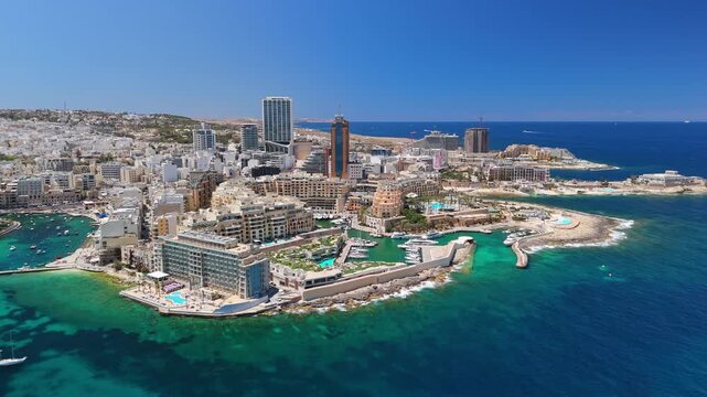 In St Julian's, Malta, an aerial view captures the marina with boats and yachts set against modern waterfront towers and hotels along the turquoise Mediterranean coastline.