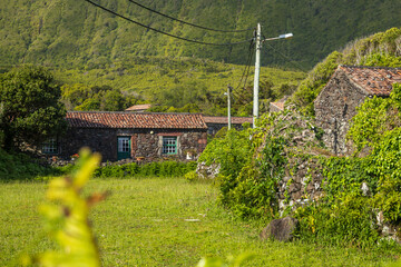 Pintoresca vista de cabañas tradicionales hechas de piedra volcánica con ventanas verdes, rodeadas de prados, muros de piedra y una gran montaña frondosa de fondo, Azores. © Maxi Perez