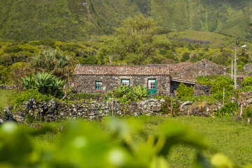 Pintoresca cabaña rústica construida con piedra volcánica y ventanas verdes, rodeada de abundante vegetación, agaves y muros de roca al pie de una frondosa montaña. © Maxi Perez