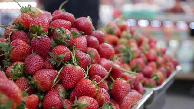 Strawberries stacked high on a retail counter forming a compact selling presentation inside a lively open public marketplace environment. Market atmosphere.