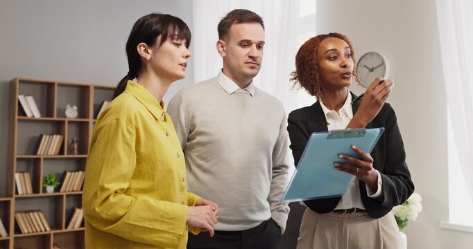 Agent guides couple during apartment property viewing. A skilled realtor offers consultation to buyers and clients, covering terms and inspection notes. Clear real estate advice concept.