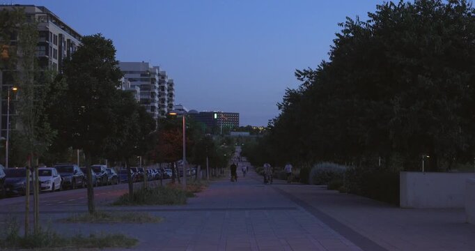 People walk along a tree-lined urban promenade at dusk. Residential buildings, parked cars and evening lights create a calm city atmosphere as daylight fades into night.