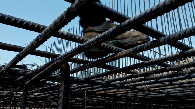 Low angle view of a construction worker walking across steel rebar reinforcement on a bridge or infrastructure site, showing civil engineering, safety equipment and structural concrete preparation.