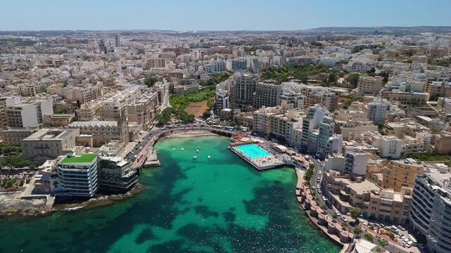 In Sliema, Malta, a bird's-eye view captures the urban coastline around the Balluta Bay with turquoise Mediterranean water, boats, and densely packed residential buildings.