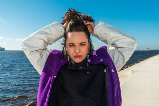 Woman adjusting hair by sea in purple varsity jacket