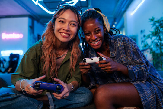 Two smiling women play video games under neon lights.