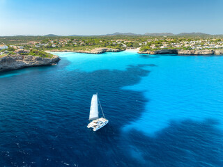 Naklejka premium Aerial view of sailing catamaran yacht in turquoise sea water bay. Sailboat. Mallorca, Spain. Luxury boat cruising in ocean cove near sandy beach, resort houses on rocky coast. Drone summer landscape