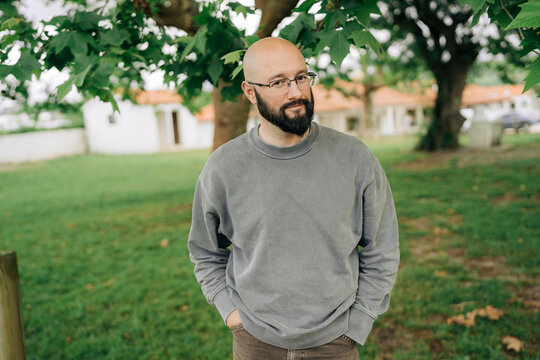 Smiling bald bearded man standing in park with hands in pockets