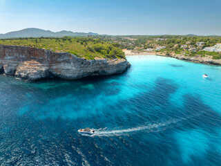 Obraz na płótnie Canvas Aerial view of motor boat near resort beach in Mallorca, Spain. Top view of Luxury speedboat cruising in turquoise sea water bay, sandy beach, hotels on high rock cliffs in summer. Drone landscape