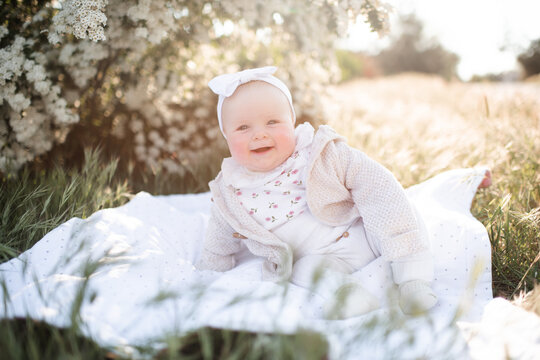 Smiling infant child under 1 year old sitting on grass lawn in sun light over blooming flowers close up. Summer season. Childhood.
