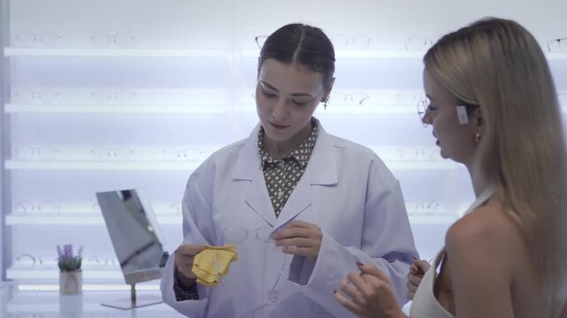 Eye care specialist in a white medical lab coat carefully adjusting trendy glasses for a blonde woman inside a modern optical shop, focusing on personalized vision care.