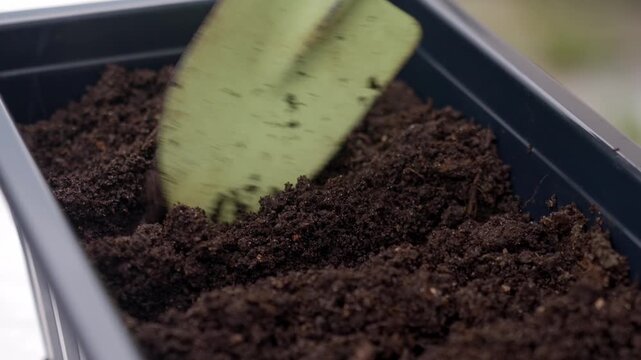 Trowel makes furrows in dark soil inside planter box for sowing seeds