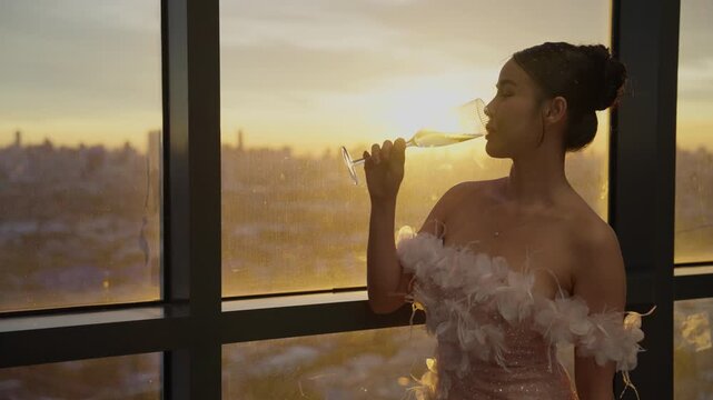 Slow motion close up of elegant Asian woman holding luxury white wine glass sipping and looking out at golden sunset city view by large window at indoor restaurant b-roll
