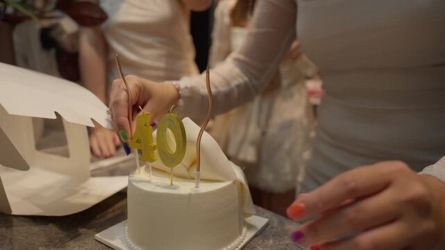 Slow motion close up of woman hand placing candle on 40th birthday white cake with group of female friends talking in soft warm light background at indoor night party celebratory b-roll