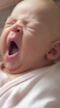 Vertical footage of a two months old baby girl is lying on a back on patterned bed linen. She is yawning so hard that she is squinting and that is looking funny and adorable