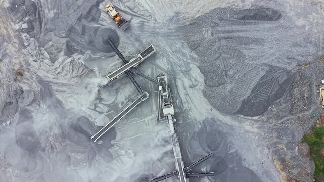 Heavy machinery operates at a gravel site next to a quarry. The equipment moves gravel and processes materials. Dust and terrain changes are visible from above, showing ongoing work.