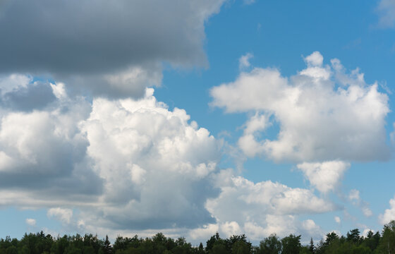Vast blue sky dominates the frame. Puffy white clouds drift lazily overhead. Gentle forest line frames the bottom edge. Sunlight illuminates cloud edges with soft glow