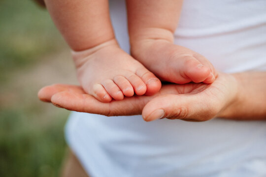 Tiny newborn baby feet resting gently on parents hands outdoors in soft sunlight. New family birth celebration, parental love care protection, organic lifestyle motherhood and fatherhood 