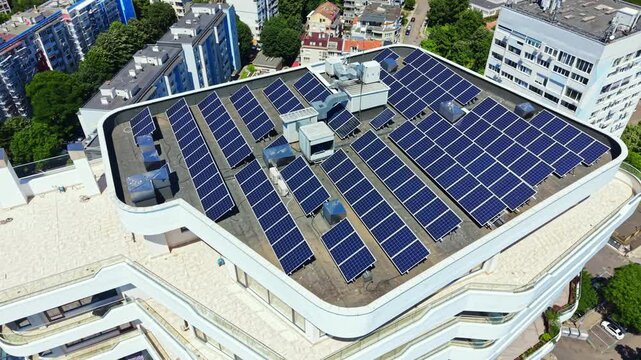 Many solar panels are arranged on the roof of a modern building in an urban area. Nearby buildings and trees can be seen in the surroundings under a clear sky.