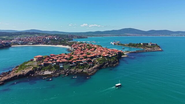 Houses with red roofs are seen on the edge of a small town by the water. A boat moves along the coast under a clear blue sky, with hills nearby.