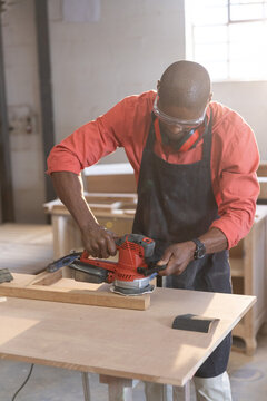 Woodworker smoothing wooden plank with electric hand sander at workshop bench, with window light