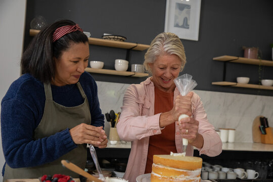 Diverse friends decorating three-layer sponge cake in home kitchen using piping bag and spatula