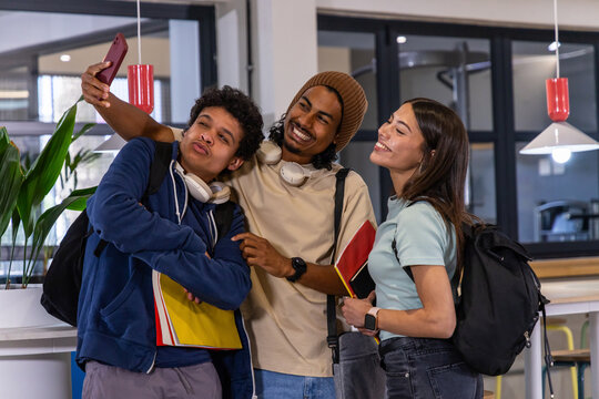 Diverse student friends posing for selfie at campus lounge holding smartphone and notebooks