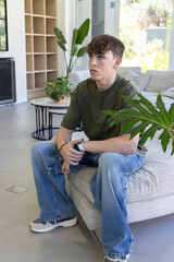 Obraz premium Adolescent male sitting on sofa in modern living room, wearing olive T-shirt with split-leaf plant