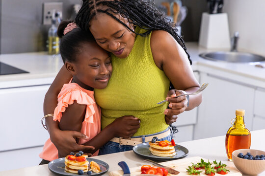 African American mother in green hugging youth daughter in peach at countertop with pancakes, fork