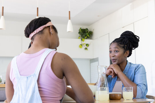 African mother and teenage daughter chatting at breakfast bar in kitchen with carafe, copy space