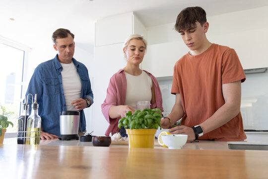 Family with parents and teenage son preparing food at kitchen island using lemon, blender, basil