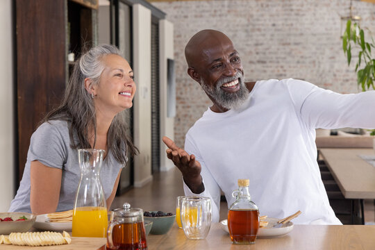 Diverse couple sitting at long wooden table in modern loft, sharing pancakes and orange juice