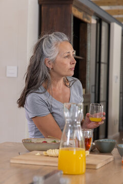 Senior woman sitting at kitchen island holding orange juice glass with carafe on board, copy space