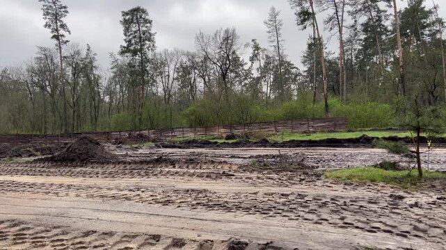 Heavy machinery is clearing and leveling the ground in a forest area. The landscape shows muddy terrain and tire tracks, indicating ongoing construction efforts.