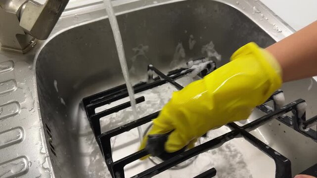Kitchen cleaning, yellow gloved hand scrubbing gas stove grate in stainless steel sink under running water, home hygiene, deep cleaning and domestic routine concept.