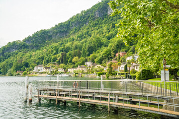 Fototapeta premium Wooden pier on Lake Lugano with village and forested mountain Caslano Switzerland