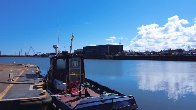 Small tugboat moored at a peaceful wharf in Matane Harbor, with crystal-clear blue water, industrial port facilities, and beautiful sky reflections.