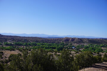 Fototapeta premium A wide angle panoramic shot of a lush green river valley oasis cutting through a rugged desert landscape, with dense trees and scattered houses nestled below rolling arid hills and a distant mountain 
