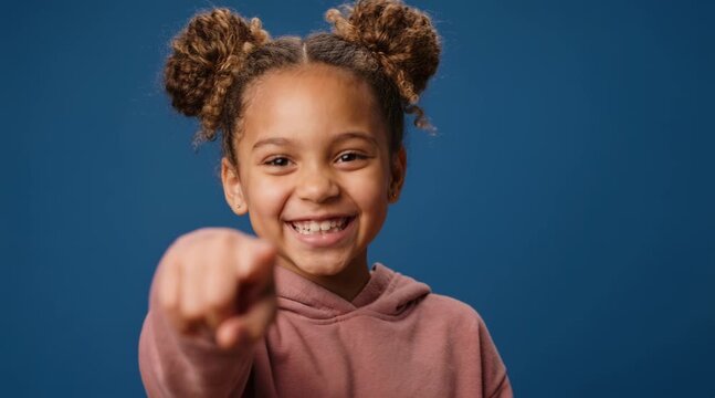 Mixed race little girl laughing and pointing at camera against solid blue studio background