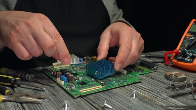 Hands of a technician installing a heatsink onto a circuit board while surrounded by tools and electronic components on a wooden workbench